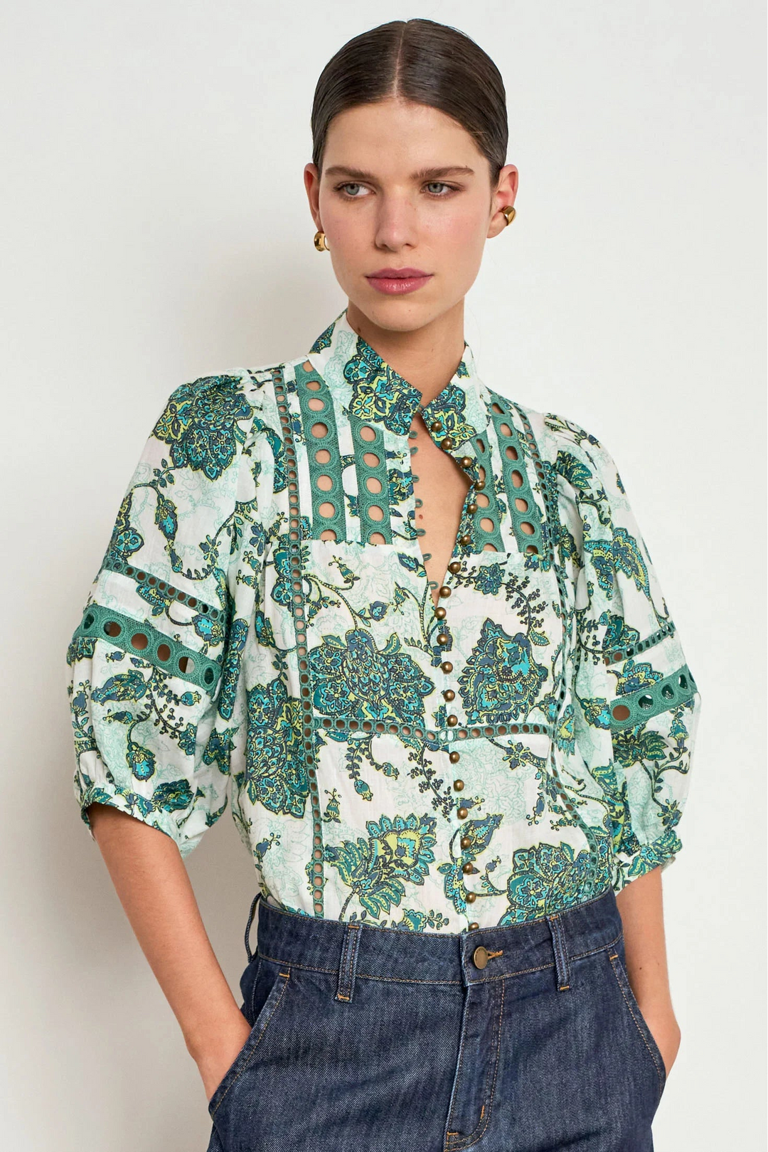 A woman modeling a short-sleeved button-up blouse with a green and teal floral paisley print on a white background, paired with dark blue jeans against a light gray studio background.