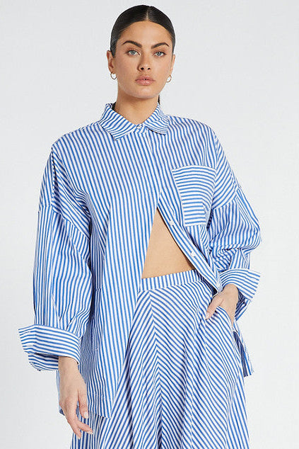 A model wearing an oversized blue and white striped shirt with a point collar, long sleeves, and side pockets. The shirt is buttoned at the top and left open at the bottom.