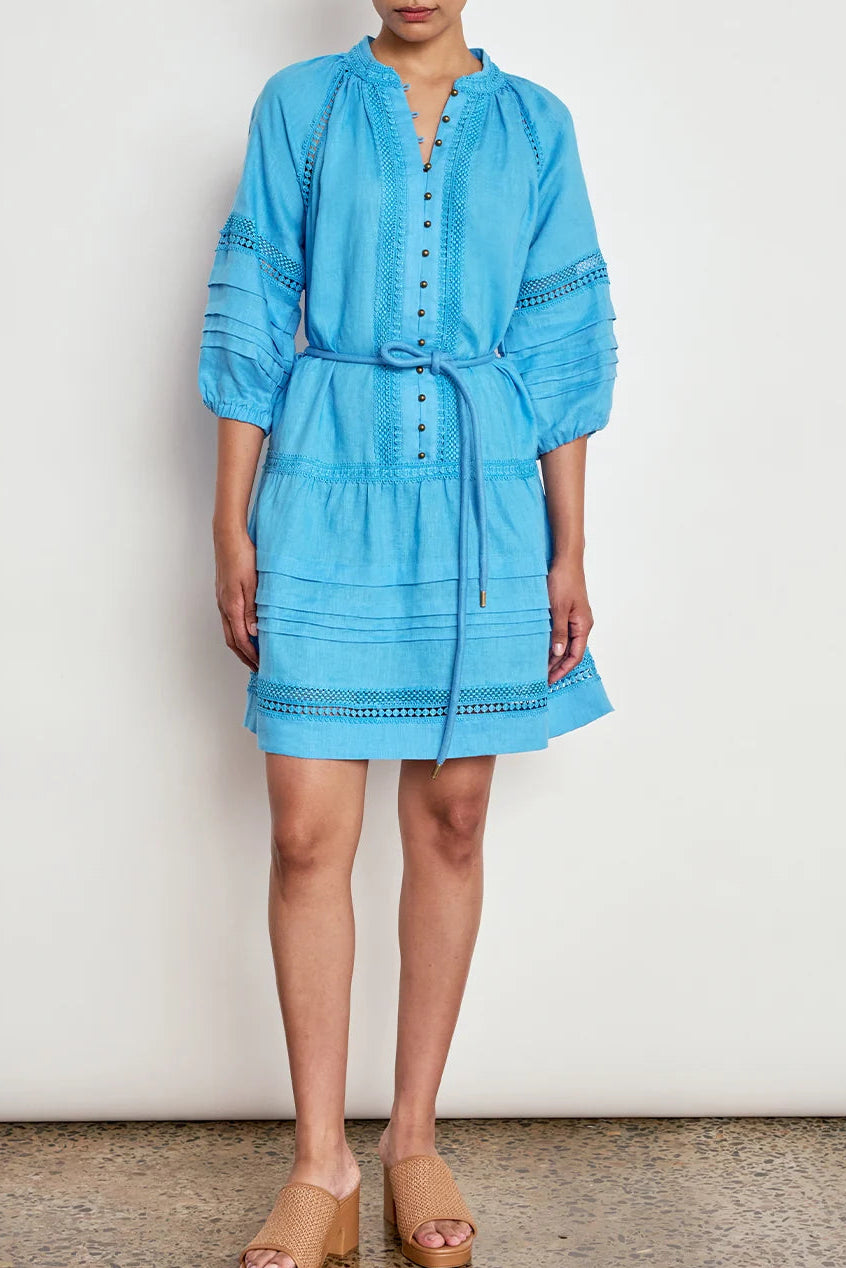 A woman modeling a light blue cotton dress with embroidered details, a button-up front, and a tie waist, paired with tan wedge sandals against a white wall.