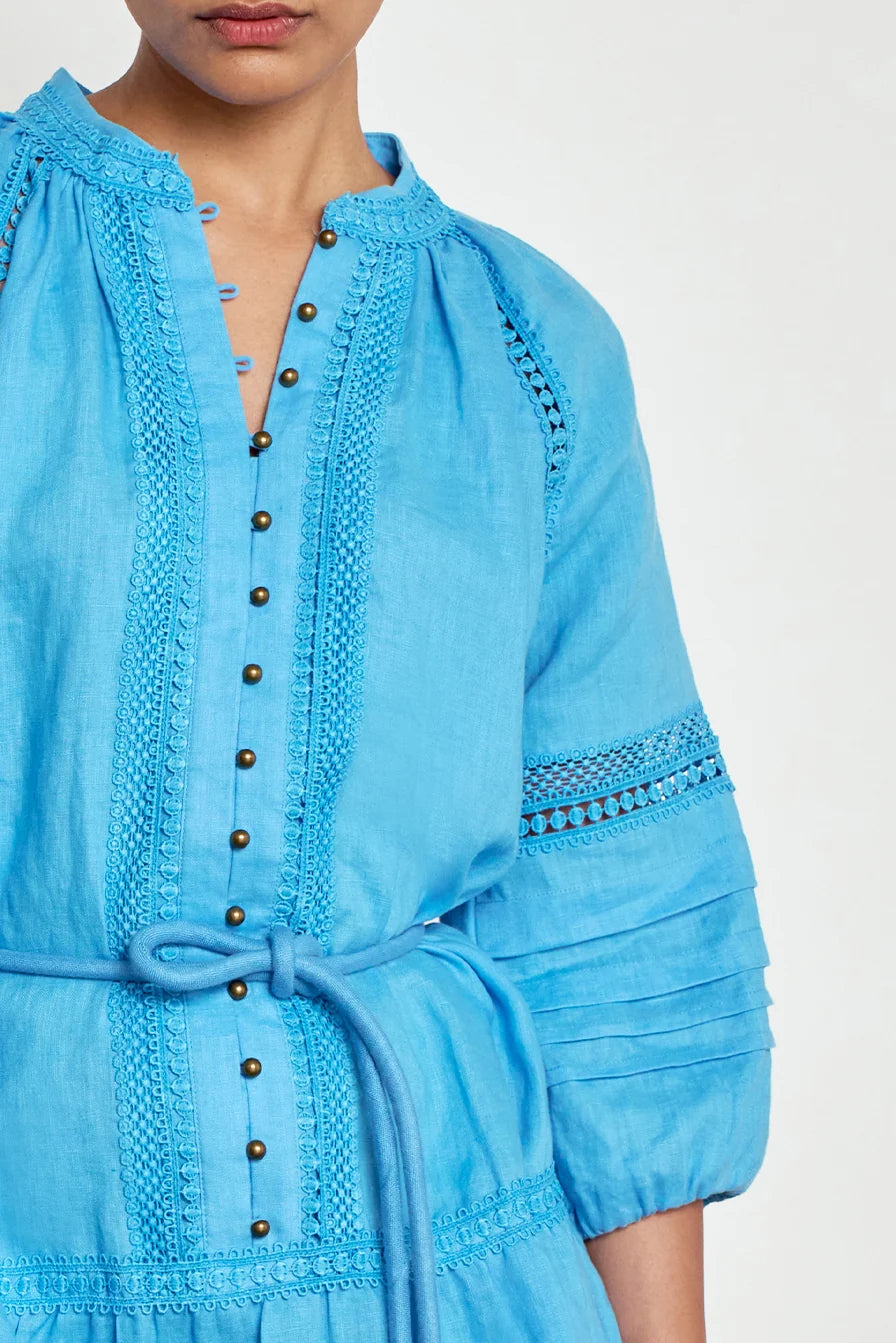 A close-up of a model wearing a light blue embroidered dress with a tie waist, button front, and detailed sleeve embroidery against a plain white background.