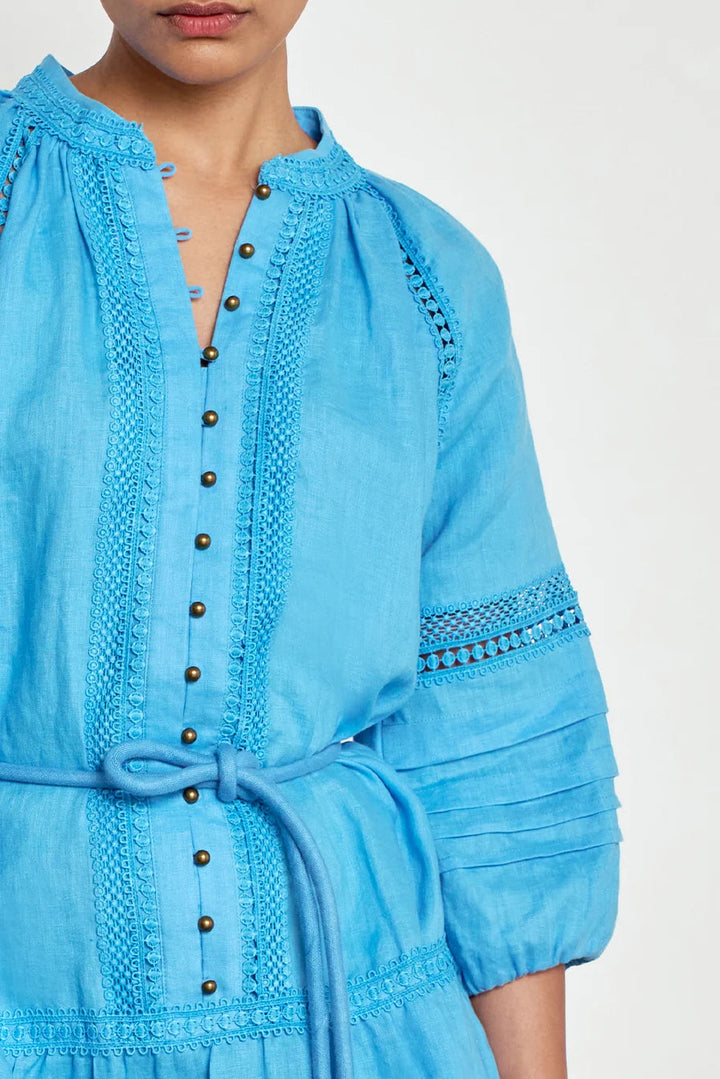 A close-up of a model wearing a light blue embroidered dress with a tie waist, button front, and detailed sleeve embroidery against a plain white background.