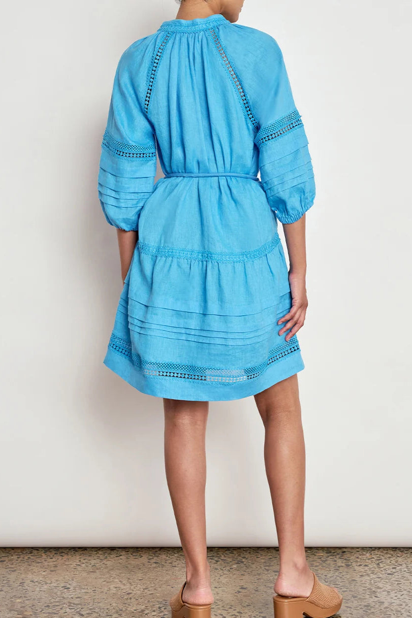A model shown from the back wearing a light blue tiered cotton dress with eyelet embroidery, paired with tan block-heel mule sandals, set against a plain off-white wall and speckled brown floor.