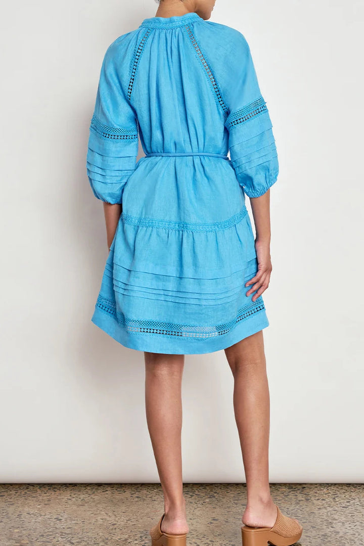 A model shown from the back wearing a light blue tiered cotton dress with eyelet embroidery, paired with tan block-heel mule sandals, set against a plain off-white wall and speckled brown floor.