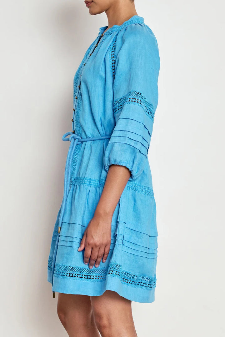 A woman wearing a light blue cotton dress with tiered ruffle details and embroidered trim, shown from a side profile against a plain off-white background.
