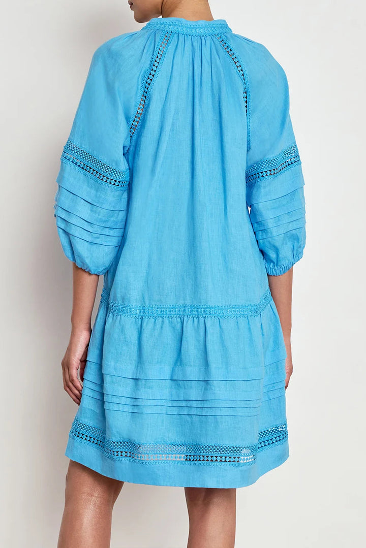 A model shown from the back wearing a bright blue tiered cotton dress with embroidered details on the sleeves and hem, set against a plain light gray background.