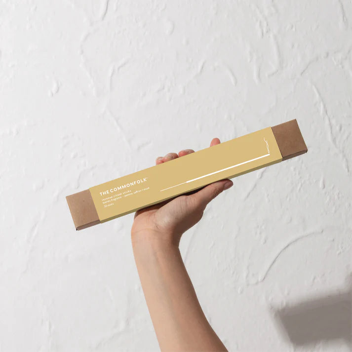 Hand holding a cylindrical product packaging with 'The Commonfolk' branding against a light textured wall.