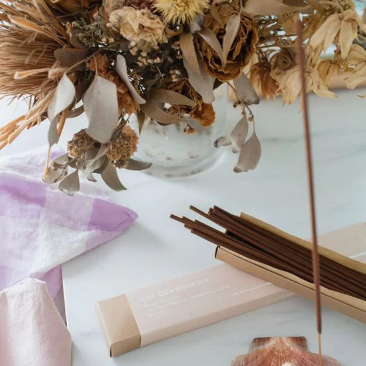 Dried flowers in a clear vase with incense sticks and packaging on a light surface.