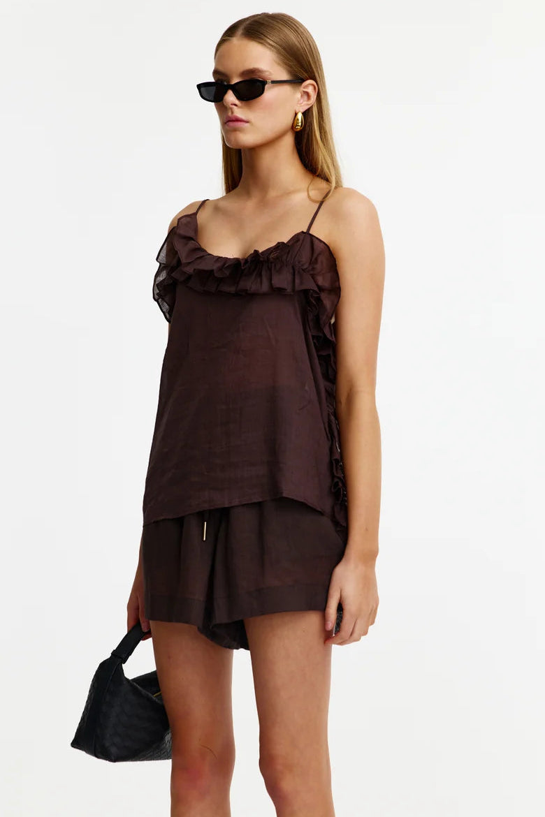 A model wearing a dark brown ruffled camisole top and matching shorts, styled with black sunglasses, gold hoop earrings, and a black quilted handbag against a plain white background.