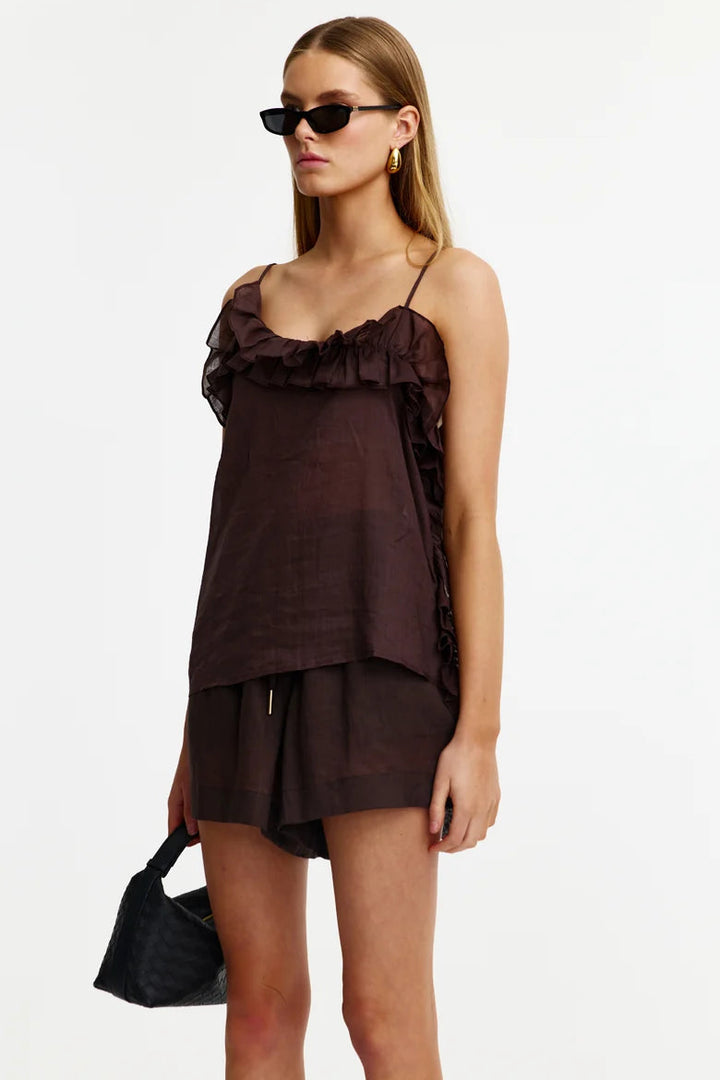 A model wearing a dark brown ruffled camisole top and matching shorts, styled with black sunglasses, gold hoop earrings, and a black quilted handbag against a plain white background.