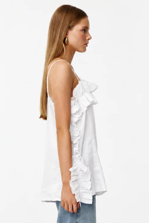 A woman stands in profile wearing a white sleeveless ruffled top and wide-leg light blue jeans against a plain white studio background.