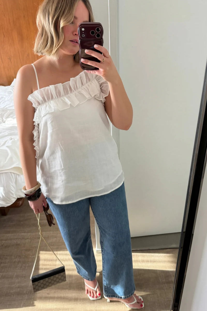 A woman takes a mirror selfie wearing a white ruffled spaghetti-strap top, blue wide-leg jeans, and pink sandals in a bedroom setting.