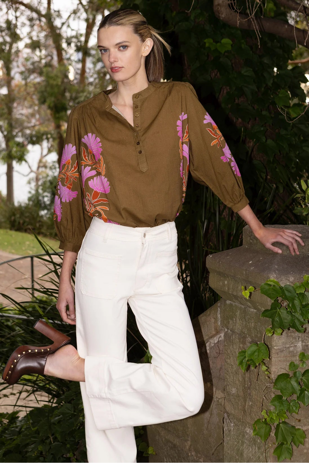 Woman wearing a brown floral blouse and white pants standing outdoors.