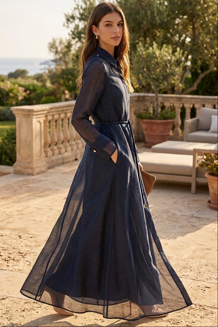 A woman wearing a long-sleeved navy blue maxi dress with a sheer overlay skirt, standing on a stone terrace with a garden and sea view in the background.