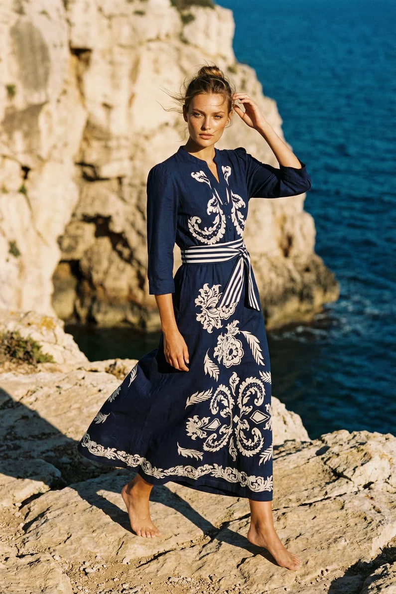 A woman stands barefoot on rocky coastal cliffs with the sea behind her, wearing a navy maxi dress with large white floral embroidery and a matching tie belt.