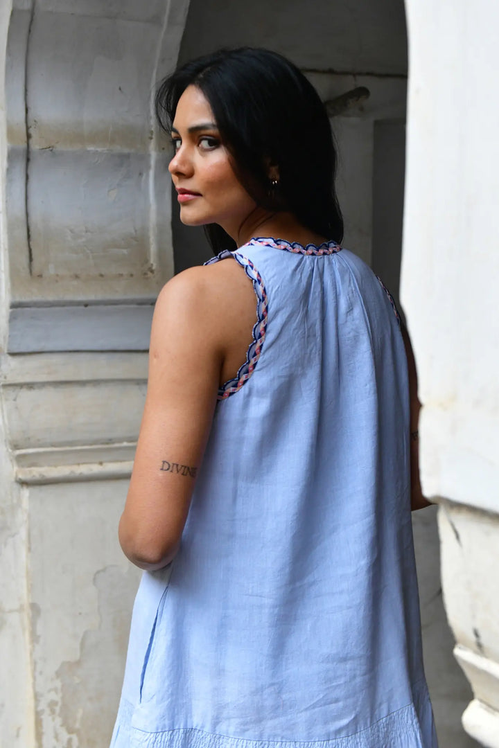 Woman wearing a light blue dress with intricate details, standing against a stone wall.