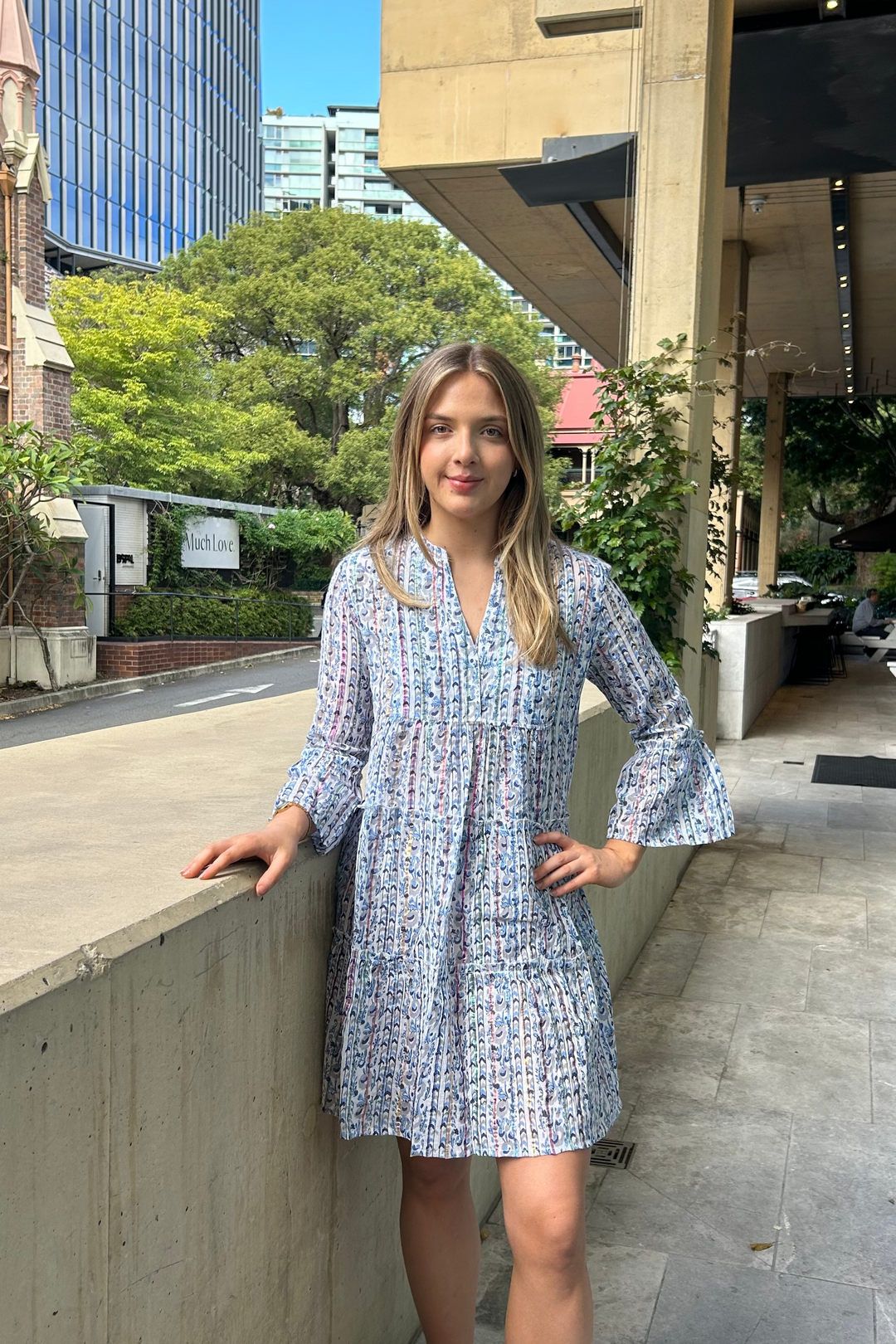 Woman in a blue patterned dress standing on a sidewalk with buildings and trees in the background