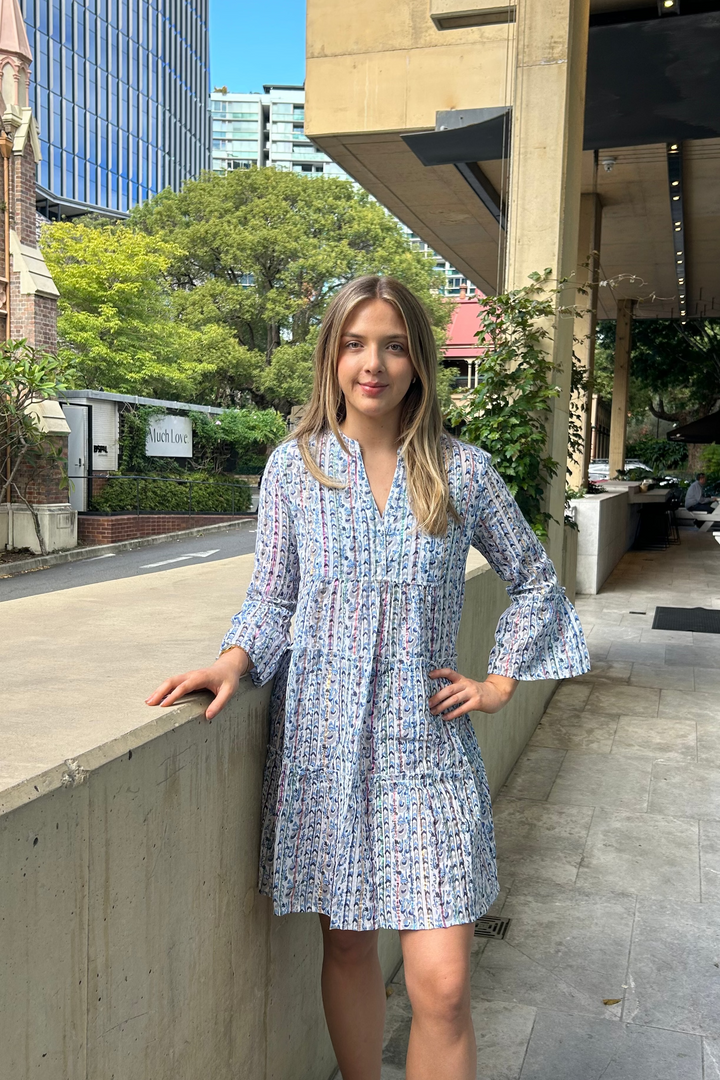 Woman in a blue patterned dress standing on a sidewalk with buildings and trees in the background