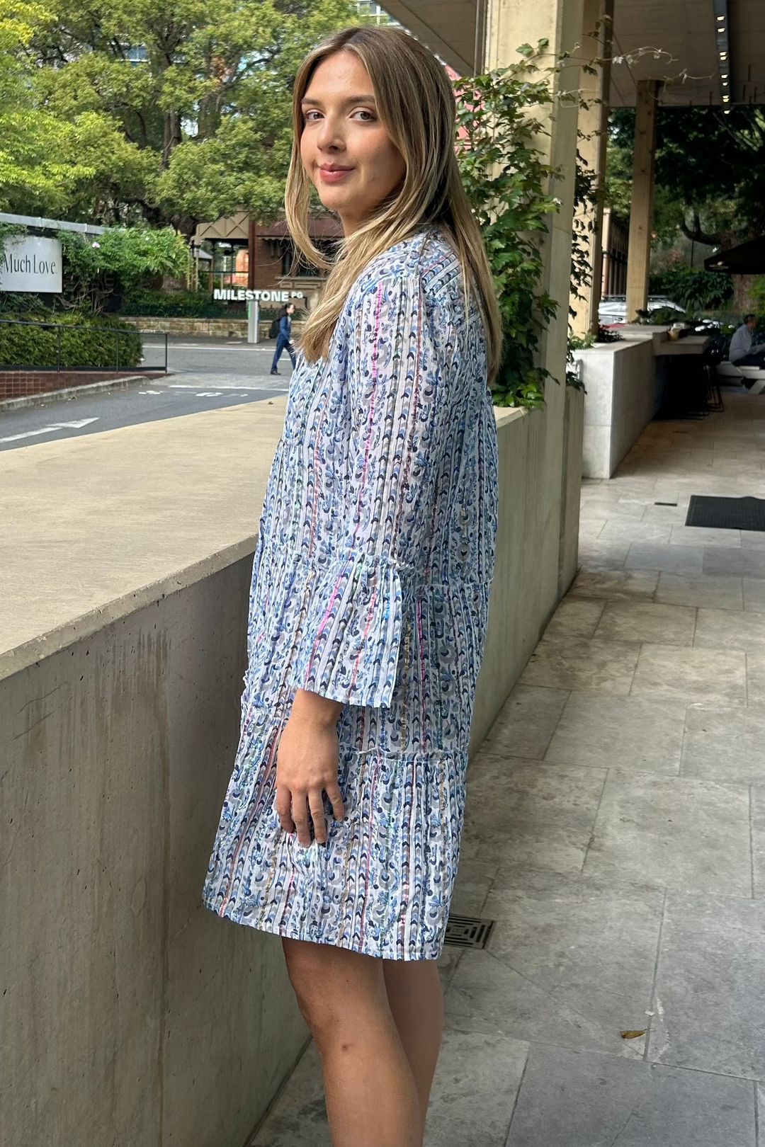 Woman in a patterned dress standing on a city street with buildings and greenery in the background