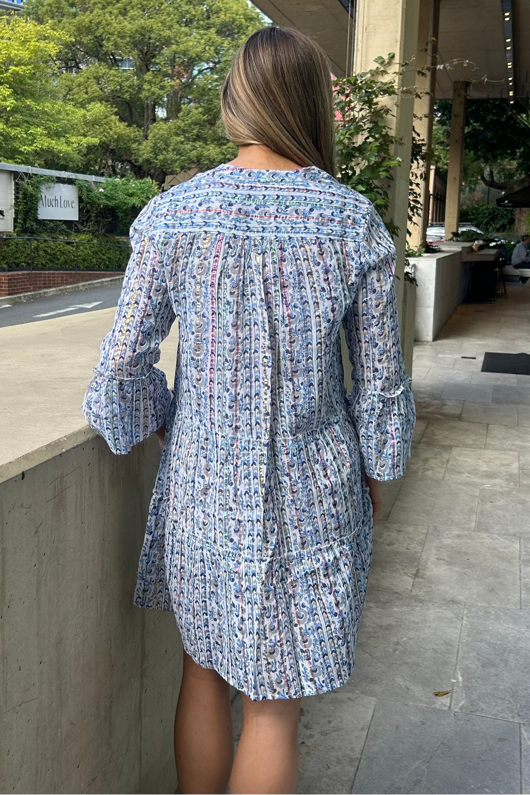Woman in a blue patterned dress walking outdoors with buildings and trees in the background