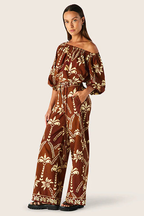 Woman wearing a brown off-shoulder jumpsuit with a palm tree pattern on a white background