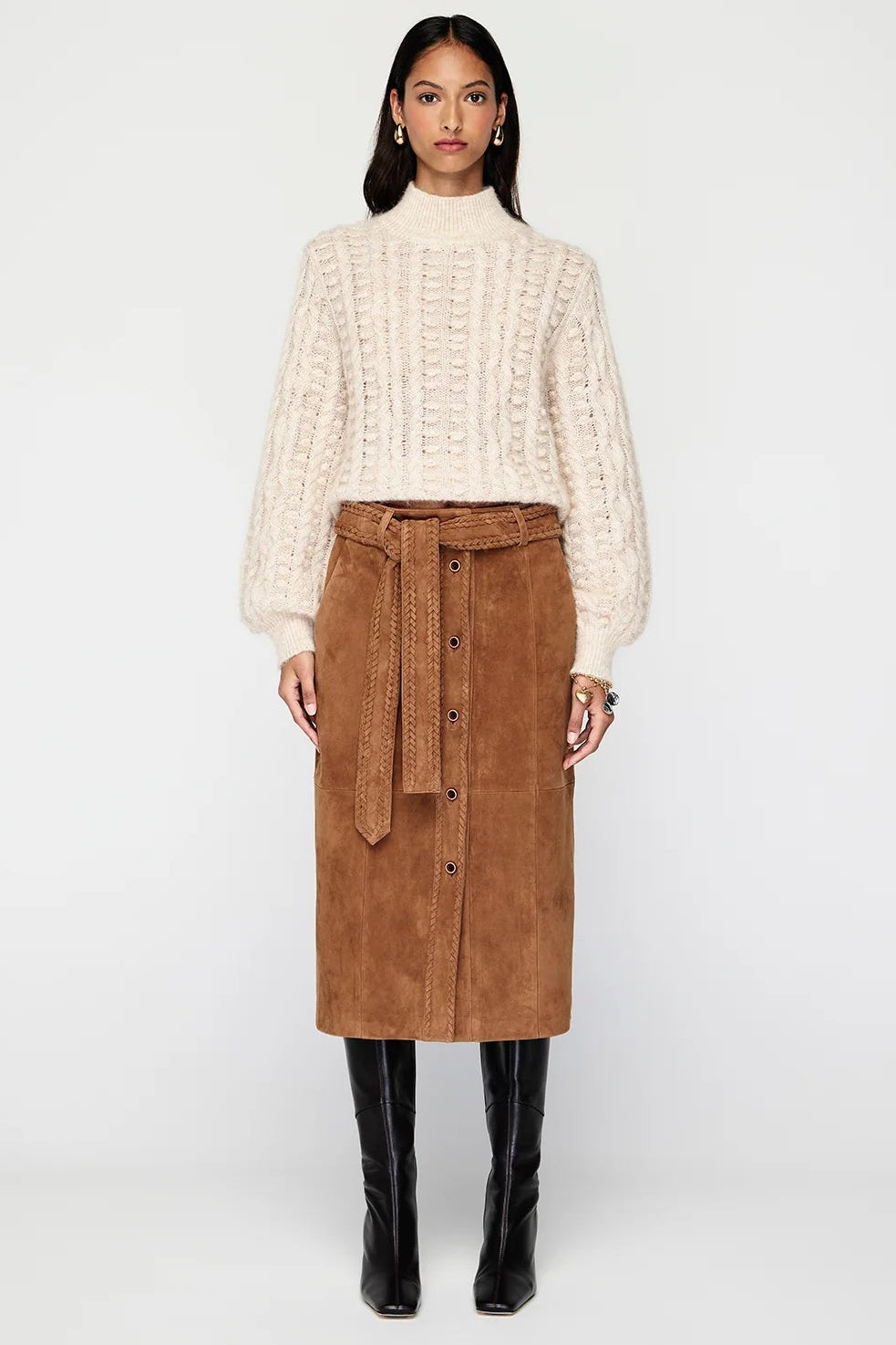 A model stands against a light gray studio background wearing a cream cable-knit mock-neck sweater, a brown suede button-front midi skirt with a matching tie belt, and black knee-high boots.