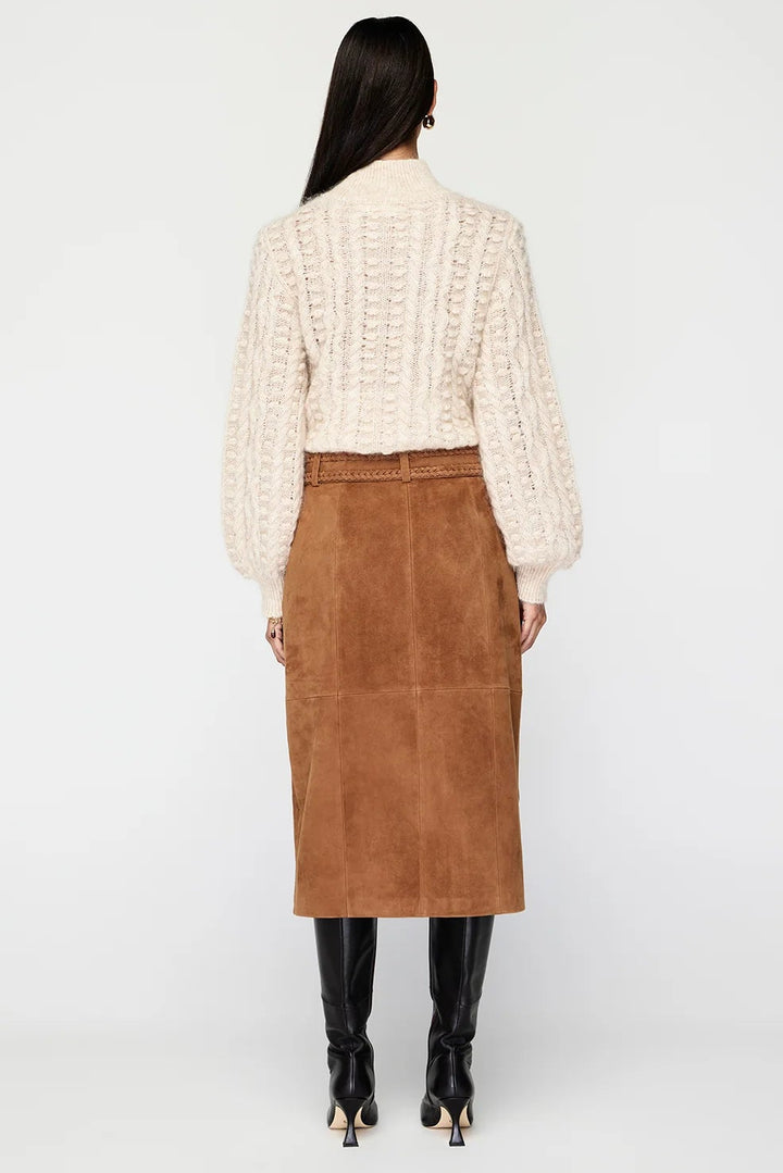 A model viewed from the back wearing a cream cable-knit cropped sweater, a brown suede midi skirt, and black knee-high boots against a light gray studio background.