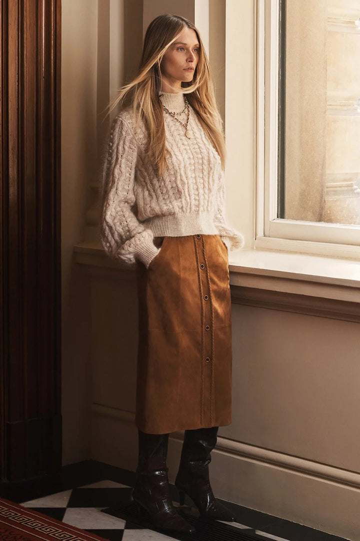 Woman in a patterned sweater and brown skirt standing by a window in a room with checkered floor.