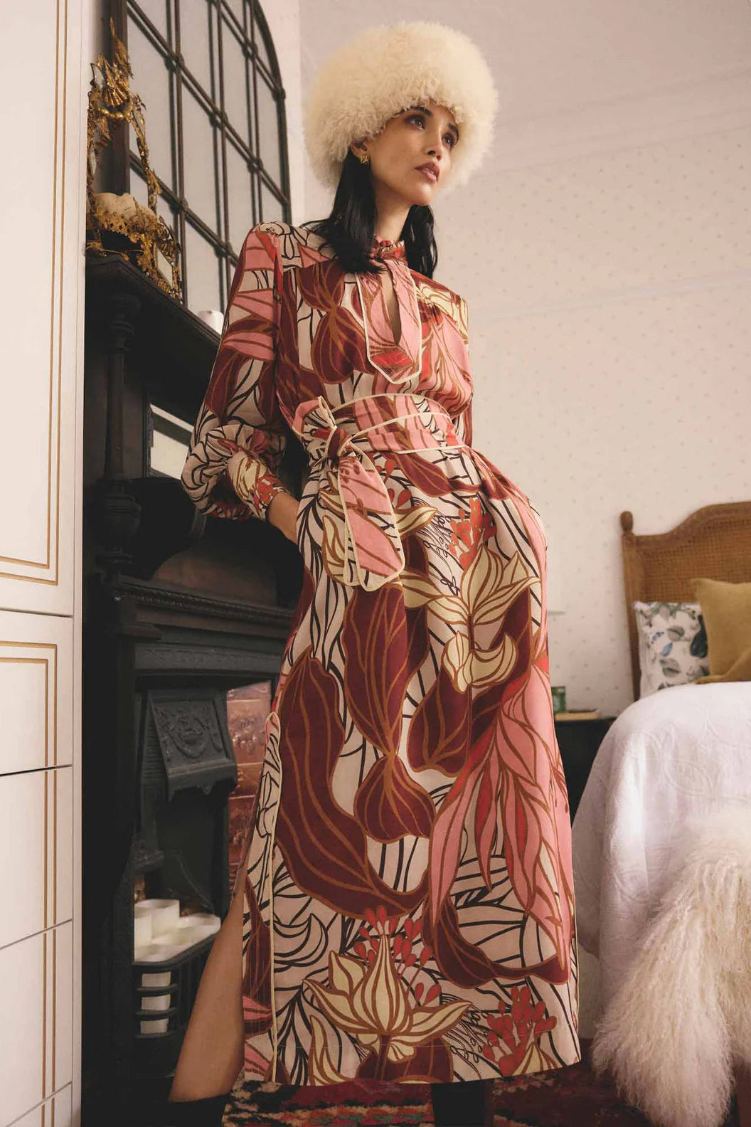 Woman in a patterned dress and fur hat standing in a room with a bed and mirror.