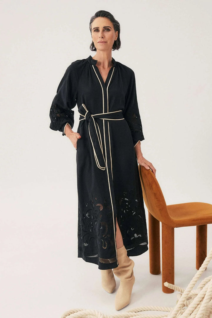 Woman wearing a black dress with white trim standing next to a wooden chair on a light background