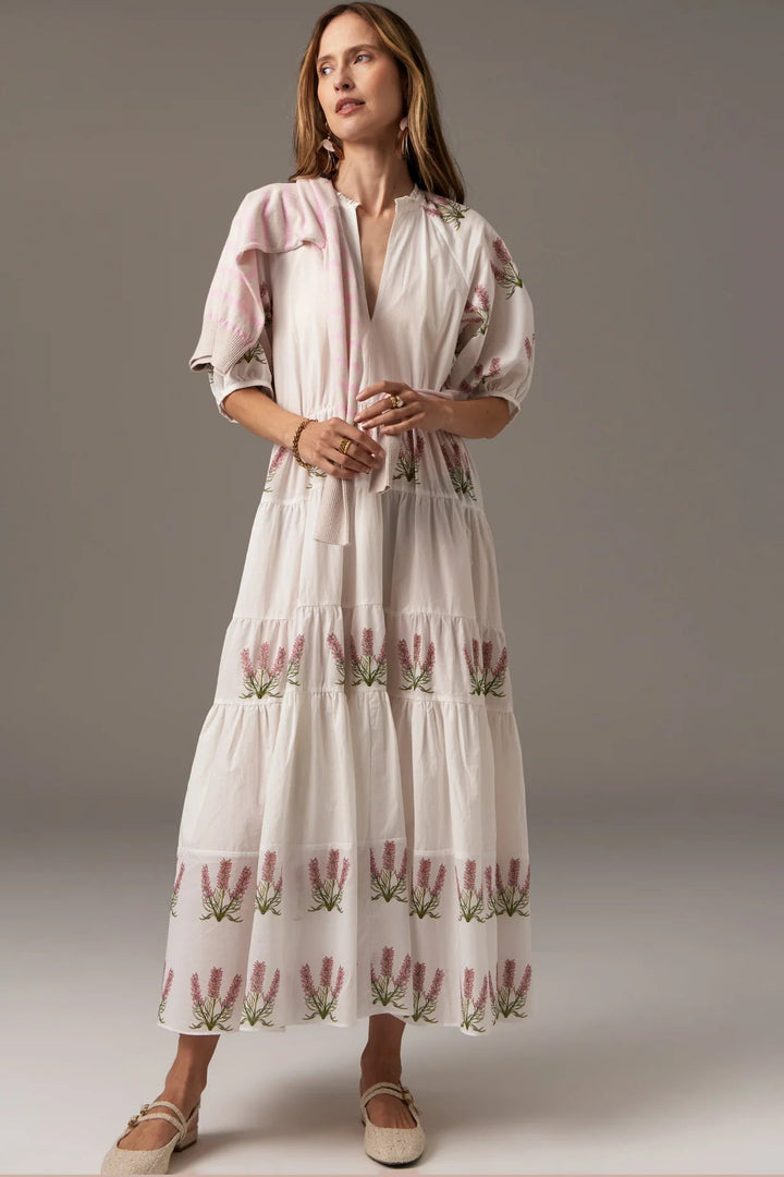 A woman standing wearing a white multicolor tiered dress with floral print and a geometric pattern, featuring a slash neckline and a tassel detail at the collar.