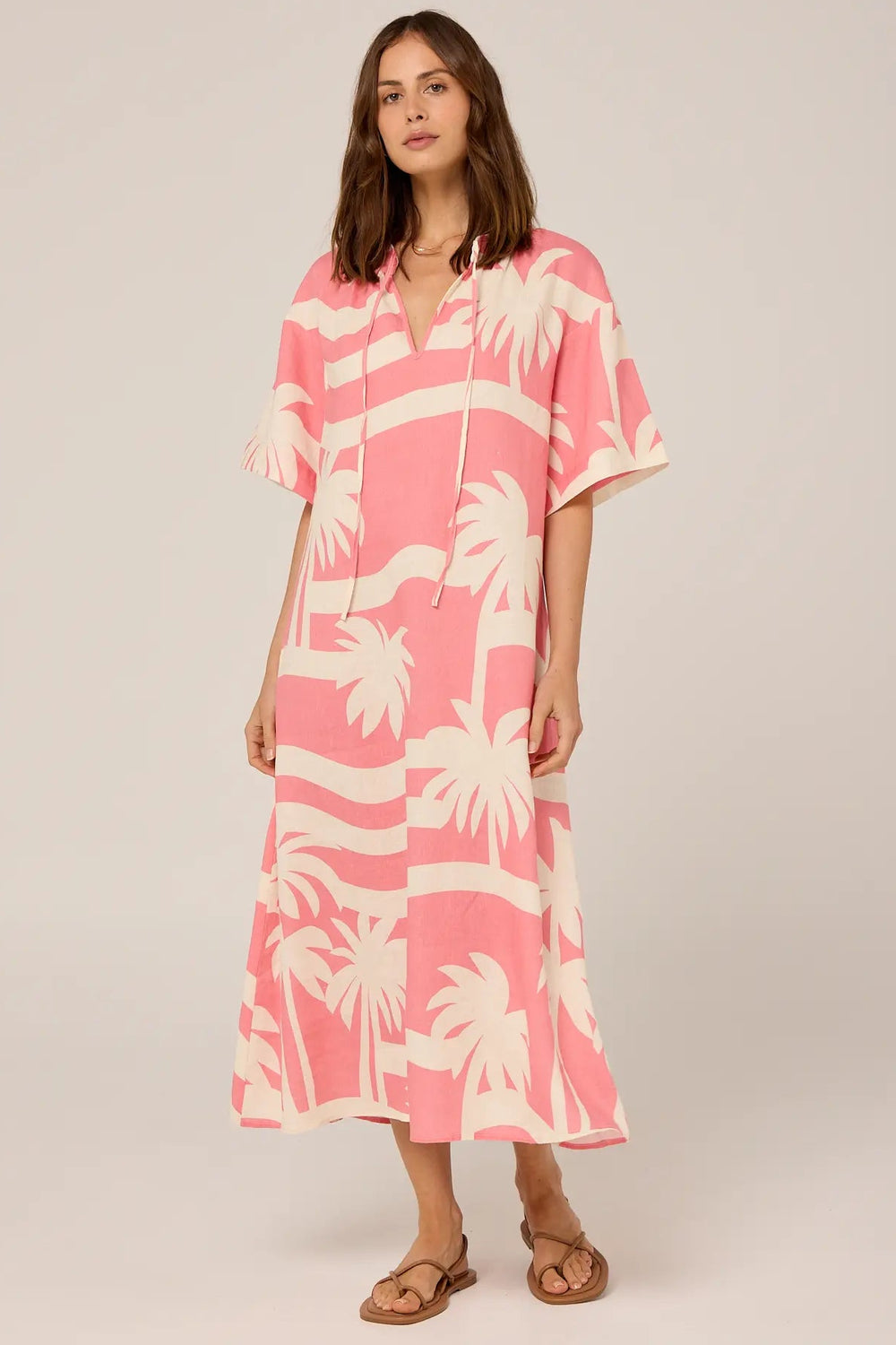 A person standing in front of a white wall with circular patterns, wearing a pink and white palm print dress with a relaxed fit and short sleeves.