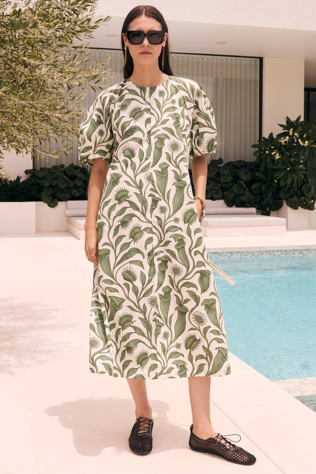 A woman wearing a green and white botanical print midi dress, black sunglasses, and black lace-up shoes stands by a swimming pool in a modern outdoor setting.