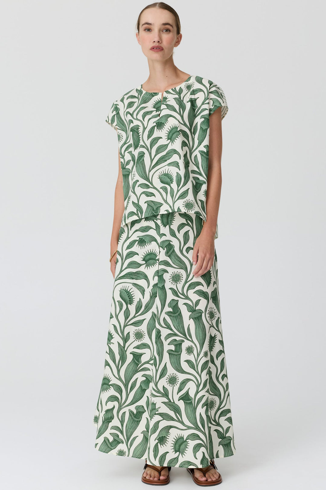 A woman modeling a matching two-piece set consisting of a short-sleeve top and a long maxi skirt, both featuring a green tropical leaf pattern on an off-white background, set against a plain light gray studio backdrop.