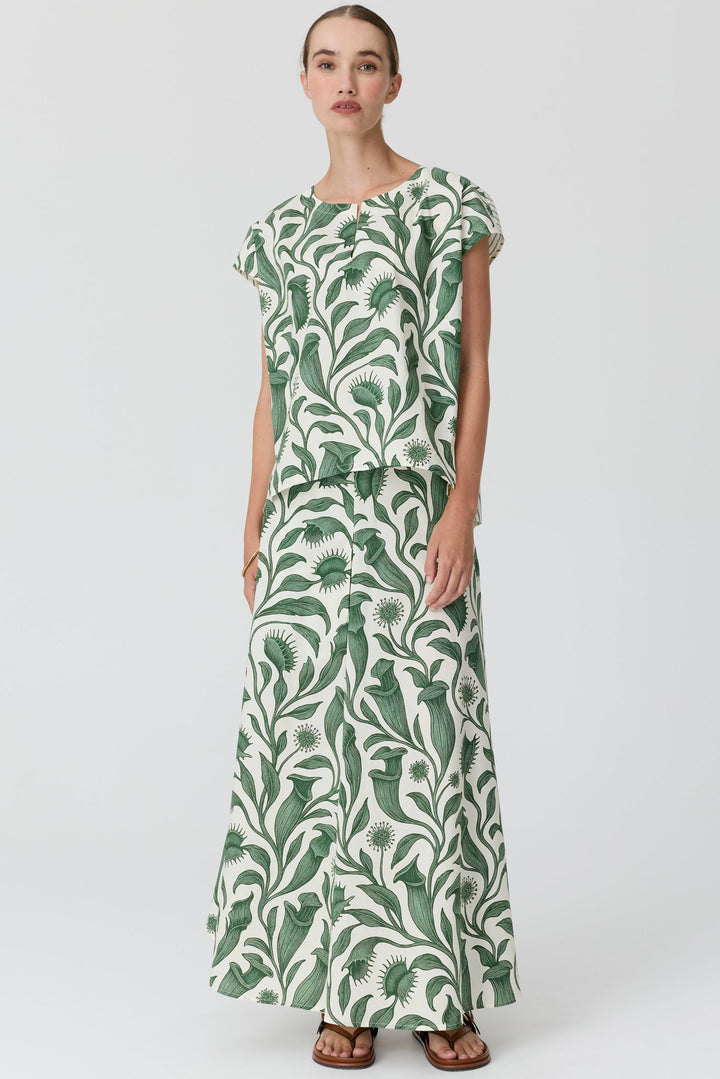 A woman modeling a matching two-piece set consisting of a short-sleeve top and a long maxi skirt, both featuring a green tropical leaf pattern on an off-white background, set against a plain light gray studio backdrop.