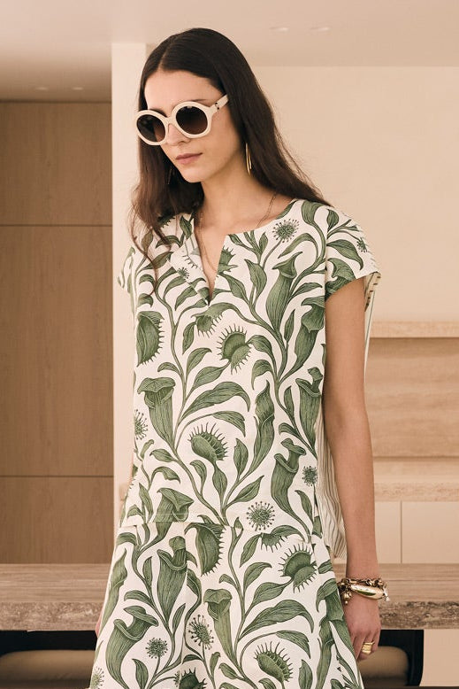 A woman wearing a green and white palm leaf print maxi dress, black sneakers, and sunglasses stands in a modern, minimalist outdoor dining area with a long stone table and dark chairs.