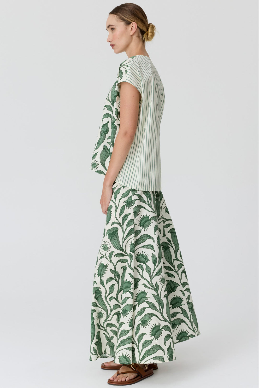 A woman shown from the side wearing a matching two-piece set with a green and white striped short-sleeve top and a long white skirt featuring a green palm leaf print, set against a plain light gray studio background.