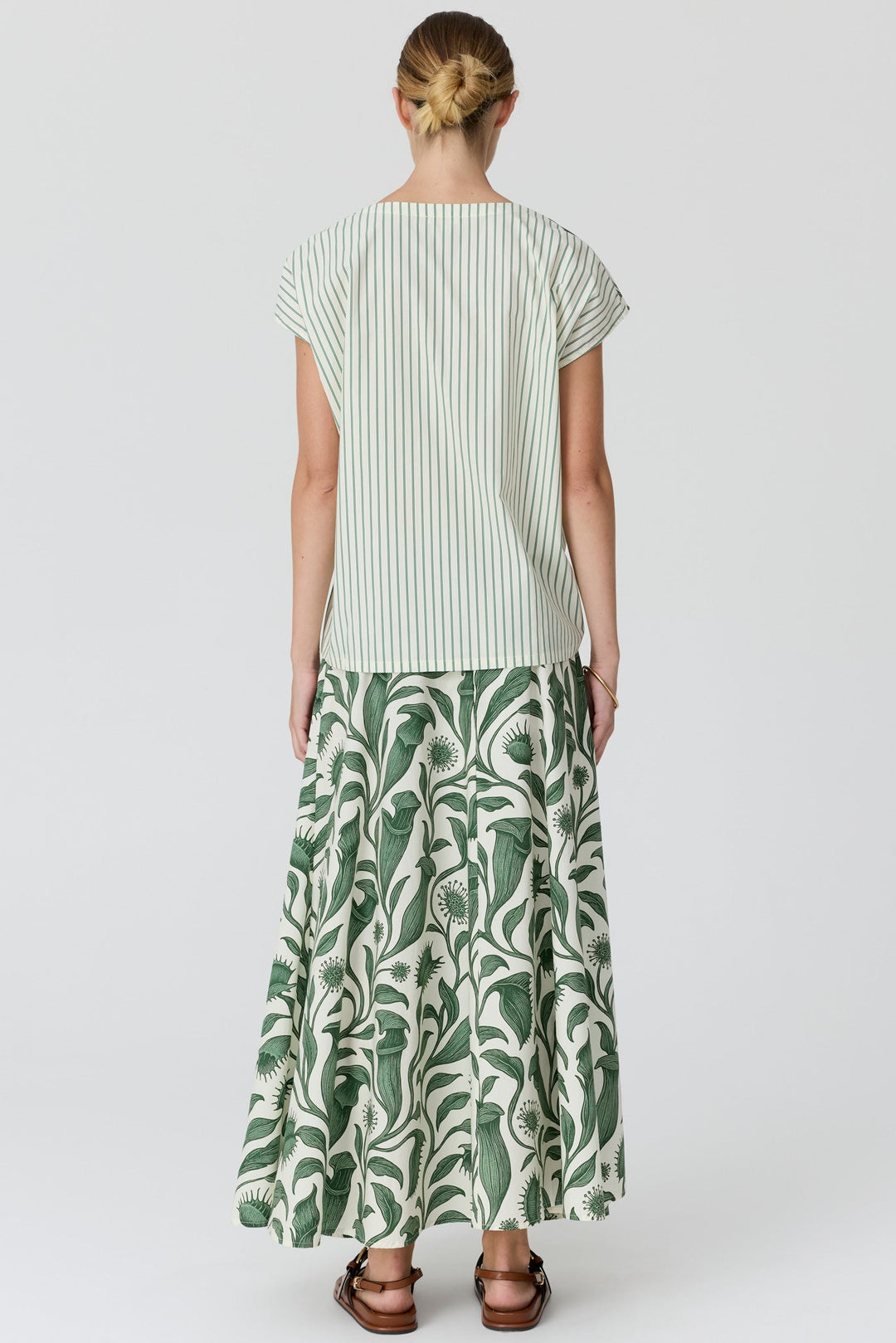 A woman viewed from the back wearing a light green and white vertically striped short-sleeve top and a matching green and white tropical leaf print maxi skirt, paired with brown sandals, against a light gray studio background.