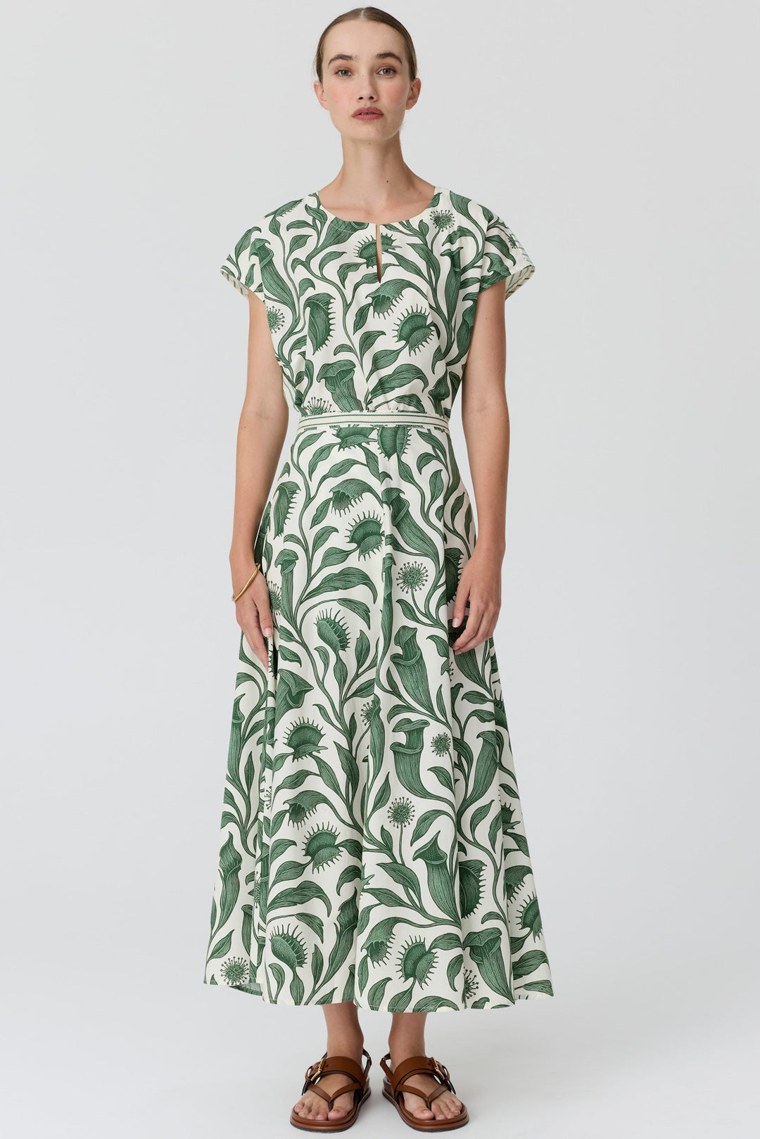 A woman modeling a long-sleeved maxi dress with a green and white botanical leaf pattern, standing against a light gray studio background.