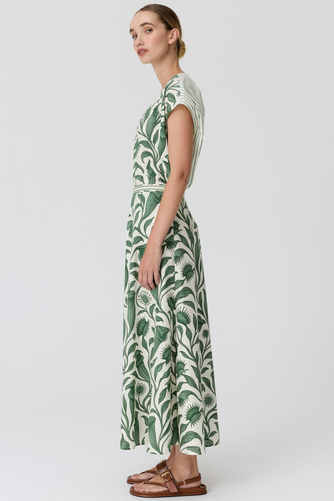 A woman modeling a sleeveless maxi dress with a green and white palm leaf pattern, shown from a side profile against a light gray studio background.