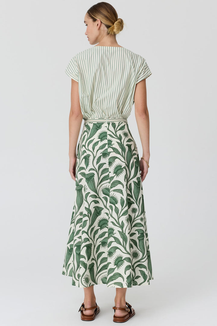 A woman shown from the back wearing a short-sleeve maxi dress with a green and off-white botanical print and a striped bodice, paired with brown sandals, against a light gray studio background.