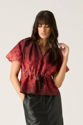A female model wearing a short raglan puff sleeve blouse with ombre floral print and elasticated waist with ribbon tie detail.