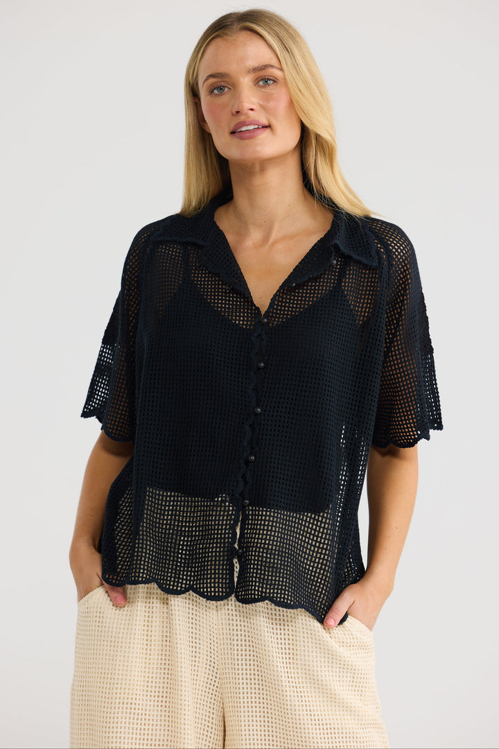 A woman modeling a black short-sleeve mesh button-up blouse with a collar and scalloped edges, paired with cream waffle-knit pants, against a light gray studio background.