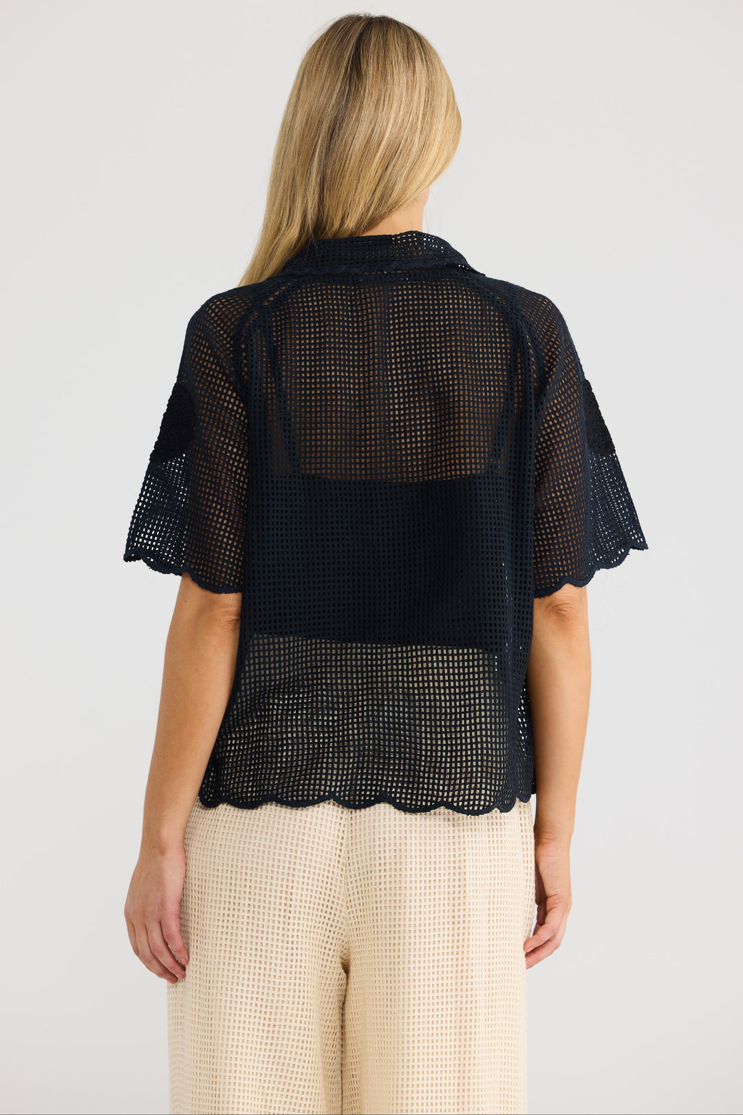 A woman viewed from the back wearing a sheer black mesh short-sleeve top with a scalloped hem, paired with light beige textured pants, against a plain light gray studio background.