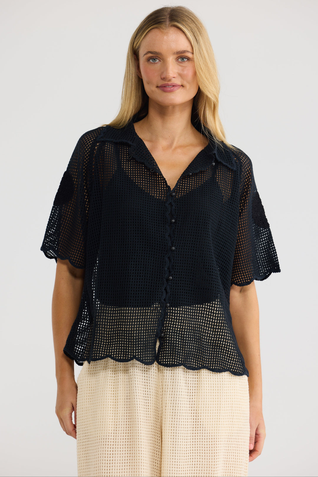 A woman modeling a black short-sleeve mesh button-up blouse with a scalloped hem, paired with cream textured pants, against a plain light gray studio background.