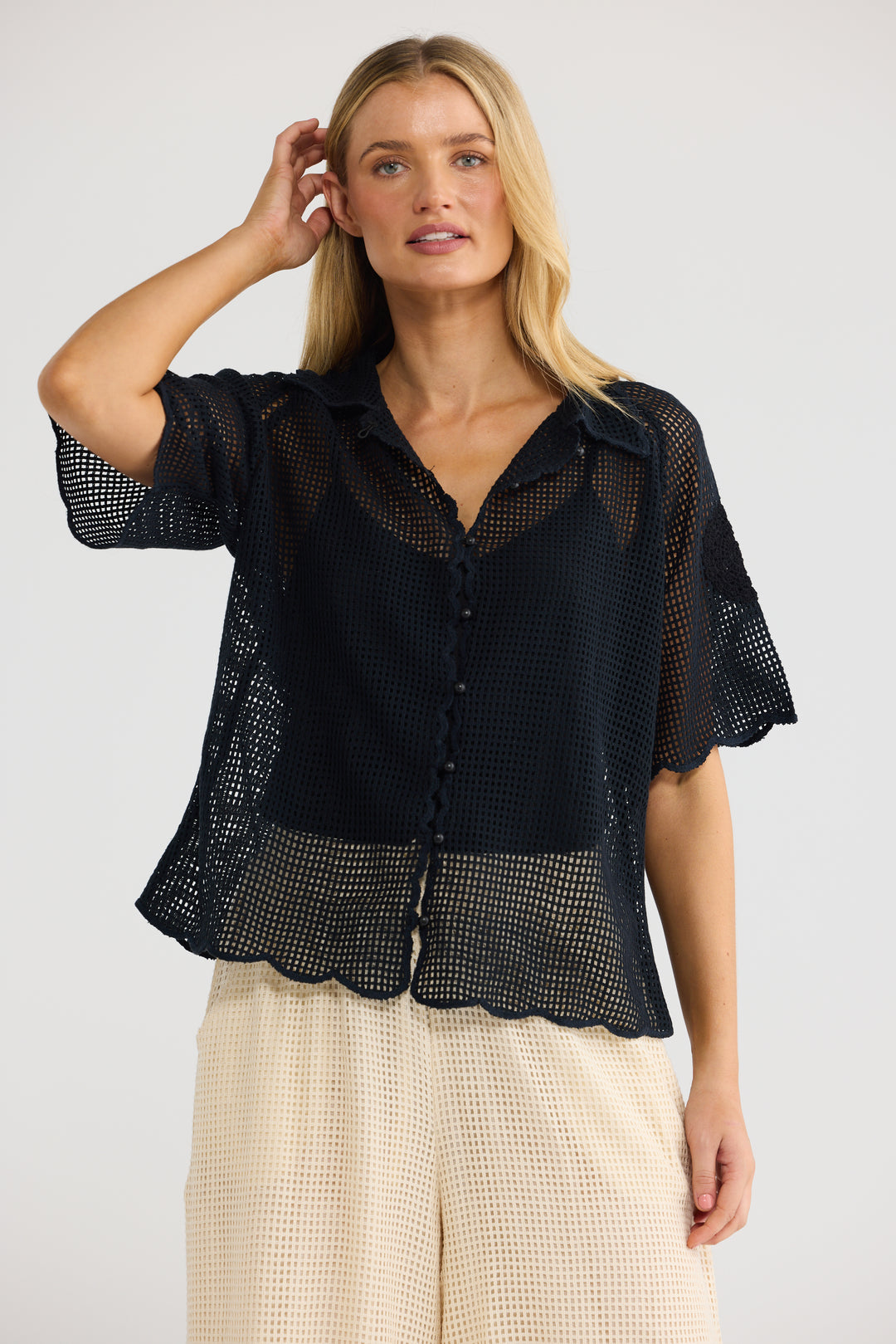 A woman modeling a black short-sleeve sheer mesh button-up blouse with a scalloped hem, paired with cream textured pants, against a light gray studio background.