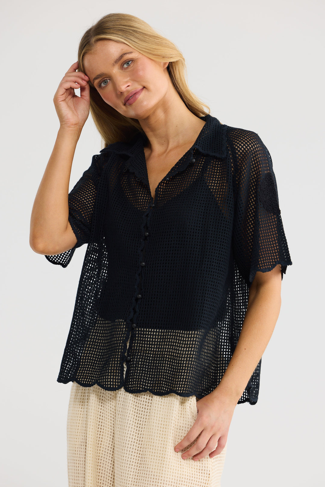 A woman modeling a black short-sleeve mesh button-up shirt with a textured open-knit pattern, paired with cream checkered trousers, against a plain light gray studio background.