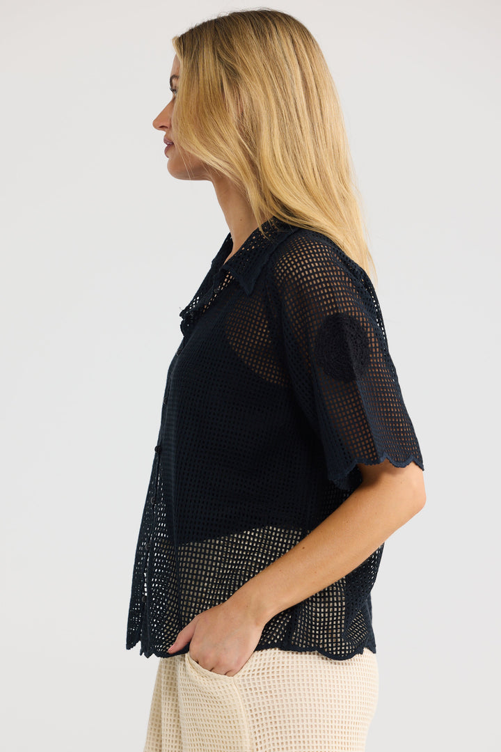 A woman shown in side profile wearing a black short-sleeve mesh blouse with scalloped edges and a textured cream knit skirt, set against a plain light gray studio background.