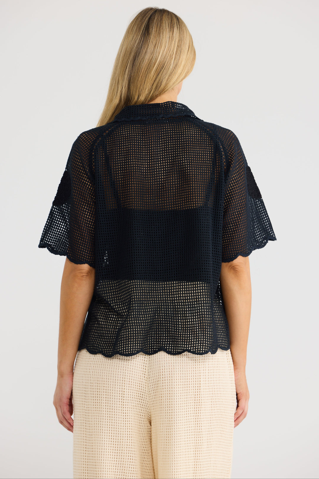 A woman viewed from the back wearing a black sheer mesh short-sleeve top with a scalloped hem and light beige textured pants, against a plain light gray studio background.