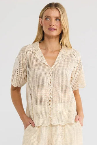 A woman modeling a cream-colored two-piece matching set consisting of a short-sleeve button-front top with a scalloped collar and wide-leg trousers, styled with brown wedge sandals against a plain white studio background.