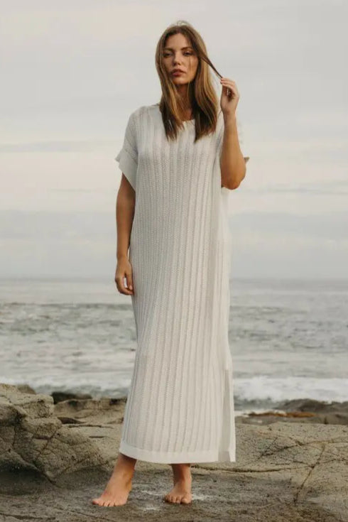 Woman in a long, light-colored dress standing on a rocky beach with ocean in the background